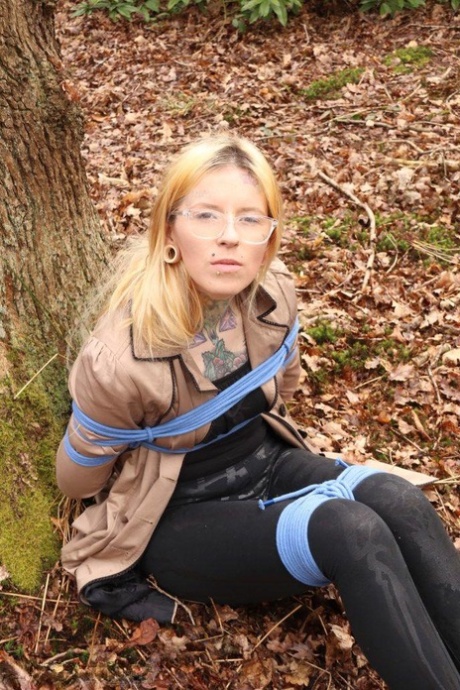 Nerdy girl is left gagged and rope bound against a tree in the forest 98562882